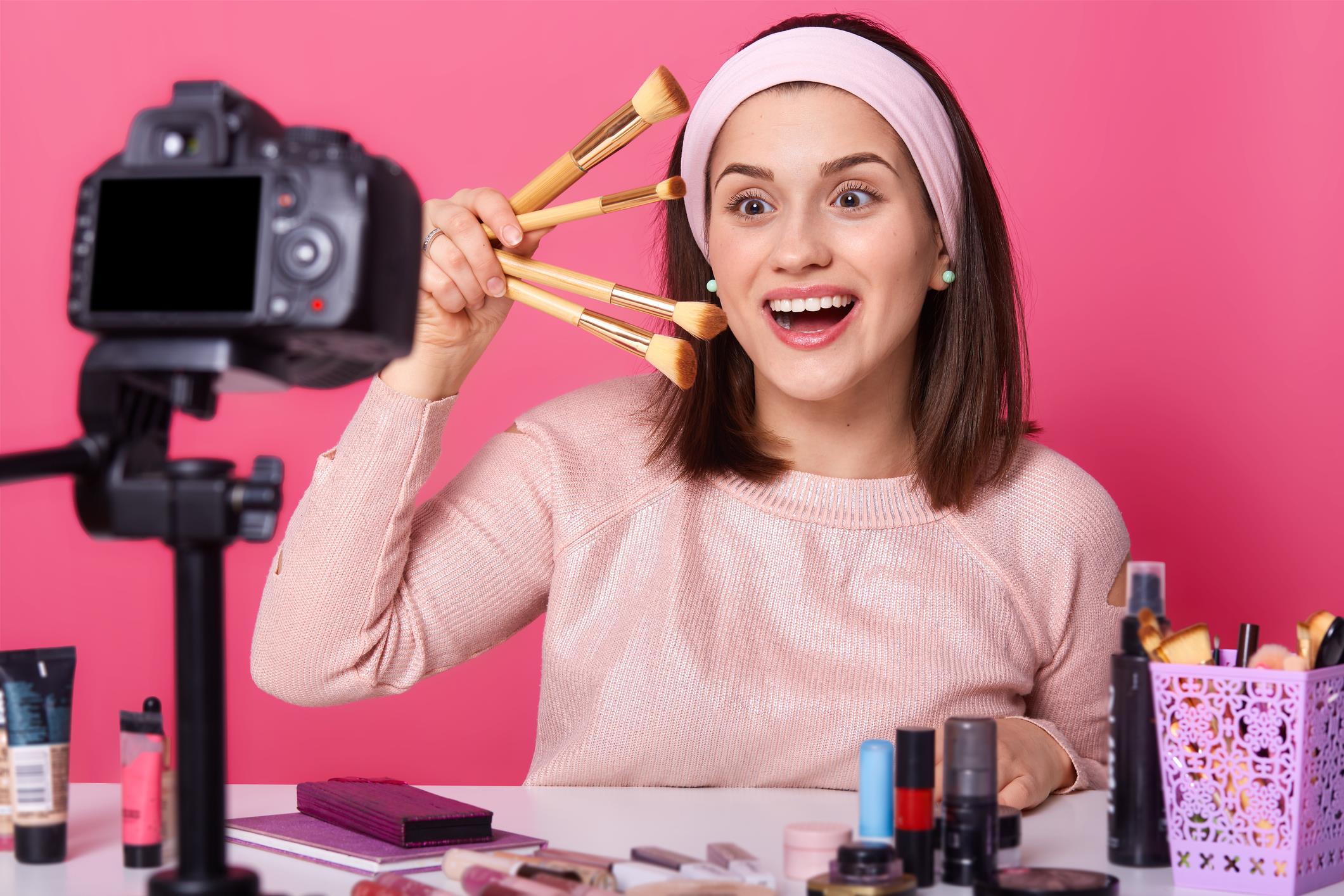 Studio shot of beautiful woman blogger stylist holds in hand different cosmetic brushes, has happy facial expression, stands with open mouth, has toothy smile. Making tutorial video for beauty vlog.